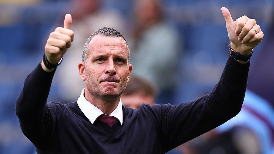 Burnley manager Nicky Hayen in a dark blue v-neck jumper, white shirt and claret tie gives them thumbs up sign to fans