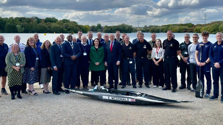 A large group of smartly dressed officials (28) assembled for the launch,  behind a black kayak in the extreme foreground at the edge of Manvers Lake, with trees and blue sky with clouds in the back ground. 