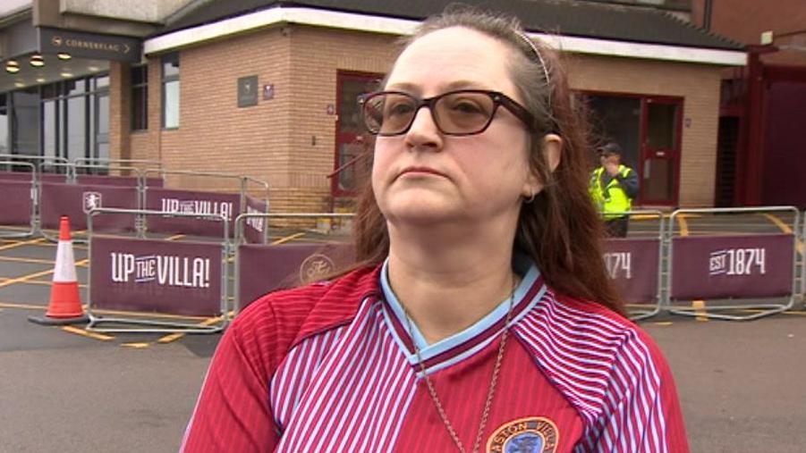 Maria Gregory - a woman with long brown hair with a headband, wearing glasses and a red Aston Villa shirt - looks to her right with a solemn expression on her face while she's being interviewed.