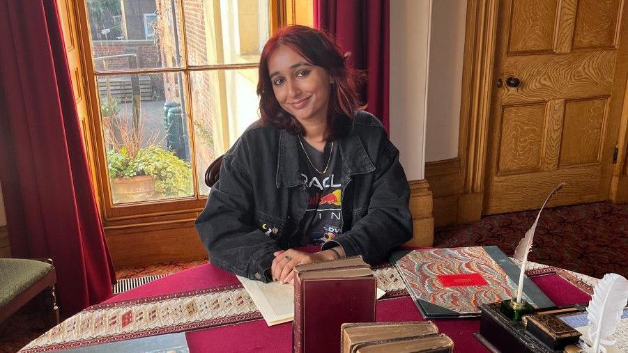 Guruleen sits at Gaskell's writing desk, which has old books and a inkstand on a red embroidered tablecloth in front of a large window