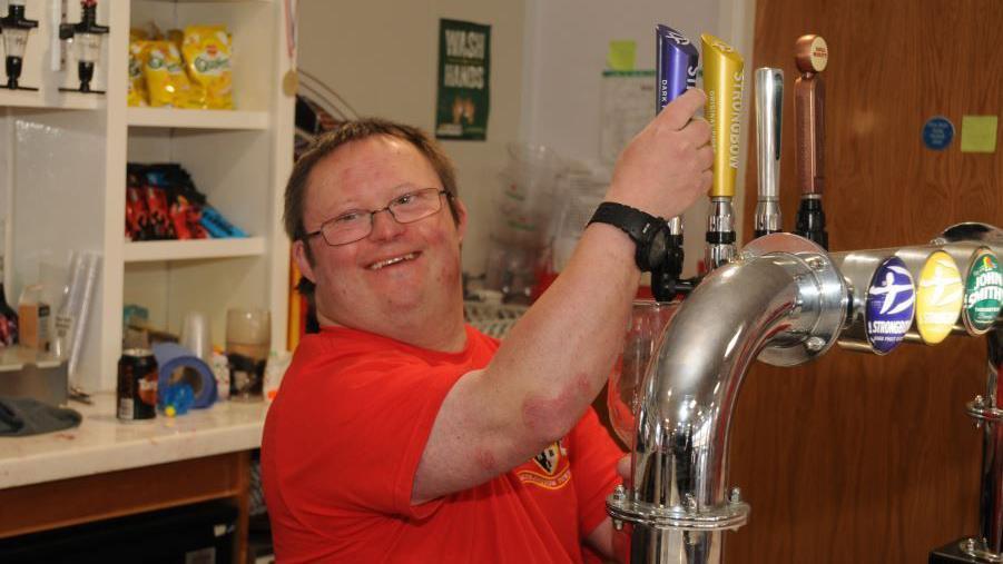 A man wearing rectangular glasses with short brown hair and a red football top on. He is pulling a pint at a bar and smiling to the side.