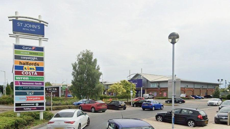 St John's Retail Park, Bedford, showing a large sign, a number of cars, buildings in the distance and a street lamp.