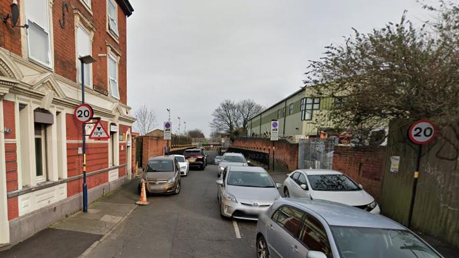 A narrow street with a red brick house on the left and what appears to be an industrial building on the right. There are several cars on the road and some parked along the pavement