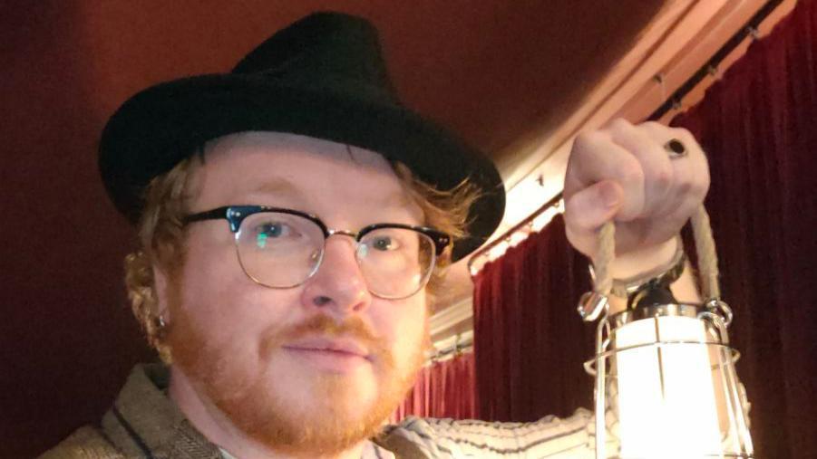 Mark holding up a lamp close to the camera, while wearing a black hat