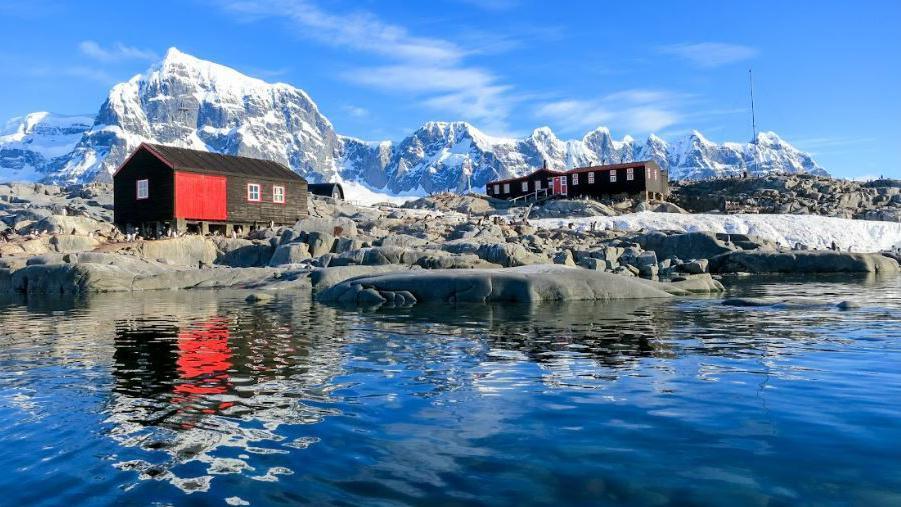 Wooden buildings set amongst snowy mountains and the sea, with clear reflections on the water's surface.