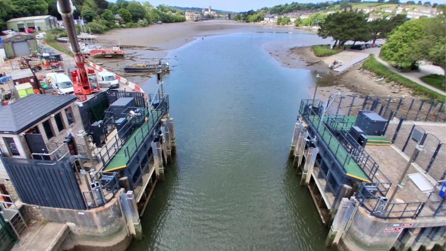 Truro tide gate, looking up towards the city of Truro. Water is in between the tidal barriers. It is low tide. Trees and green spaces are on land on either side of the water. Properties are in the distance.