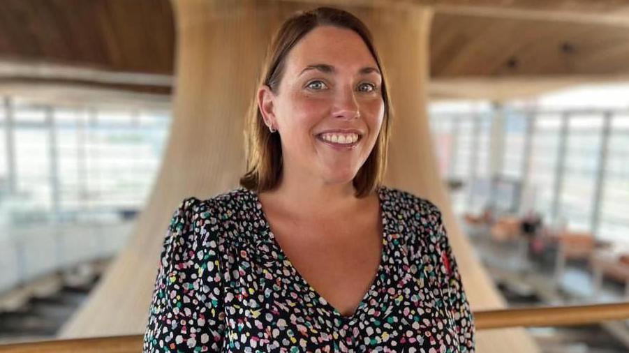 The image shows Heledd Fychan standing in the Senedd looking directly at the camera and smiling. She is wearing a colourful dress. The Senedd's funnel is behind her. It is a head and shoulders shot of her.