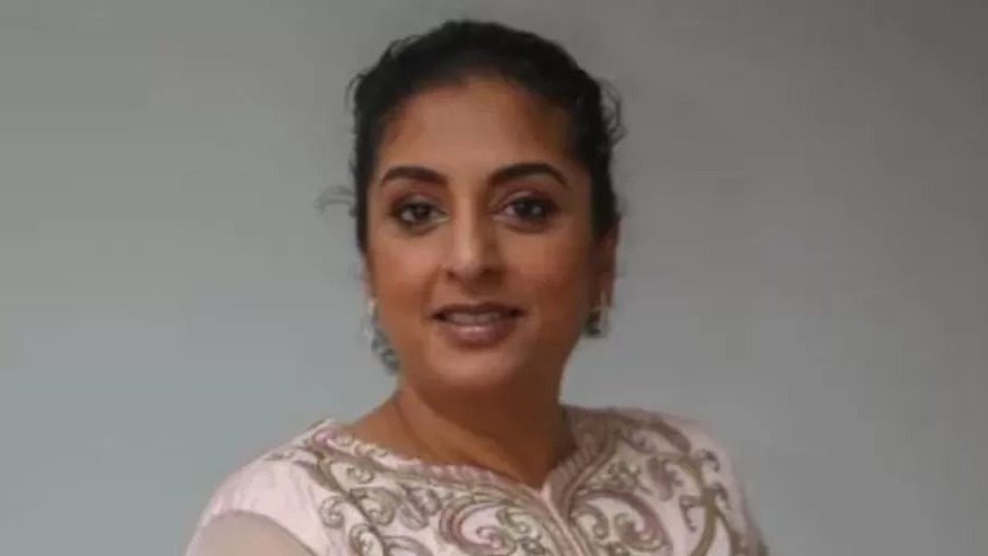 Angela Mittal is pictured with a pink dress and has her black hair tied back and is wearing dangling earrings. The background behind her is grey 