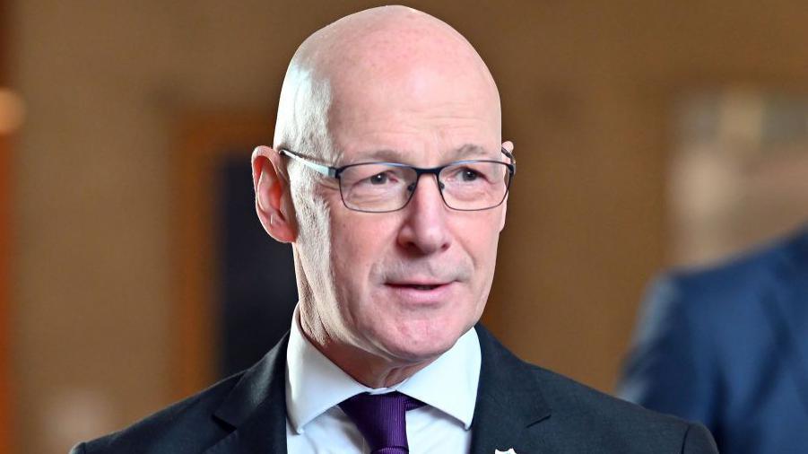 John Swinney, a bald man who is wearing glasses. He has on a suit with a purple tie. His expression is minimal with a slight smirk. The corridors of Holyrood are out of focus in the background.