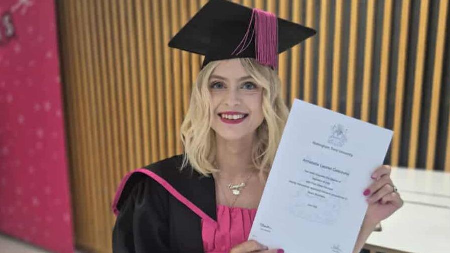 A woman wearing a black and pink university gown and cap at her graduation ceremony. She is holding her degree certificated, a white A4-sized piece of paper - in front of a black and gold background. 