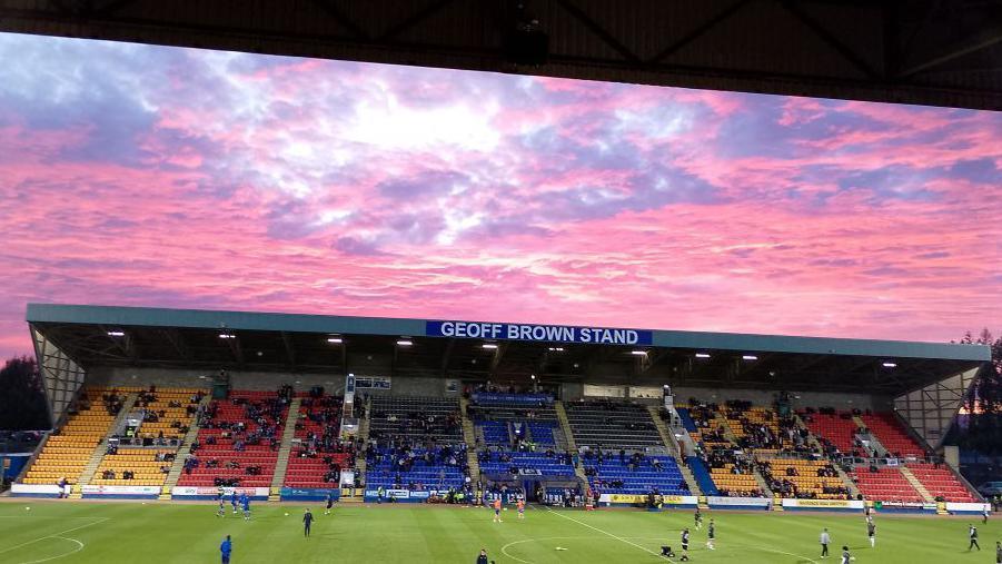 Pink sunset sky over McDiarmid park football pitch, with yellow, red and blue seats in the stands, with fans in place and players warming up on the green pitch.