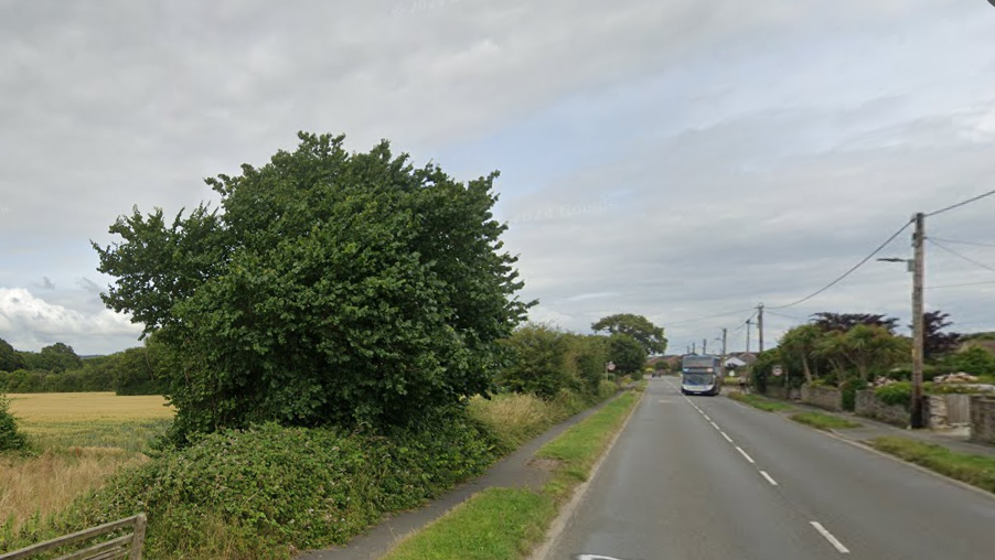 A street view of the B3233. There is a double decker bus in the distance on the right and houses on the right. On the left is a field, bushes and trees.