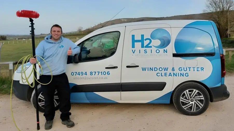 Jason Knight stands in front of a van holding onto his window cleaning pole. The van has the logo for his company H2O Vision and has blue and white graphics.