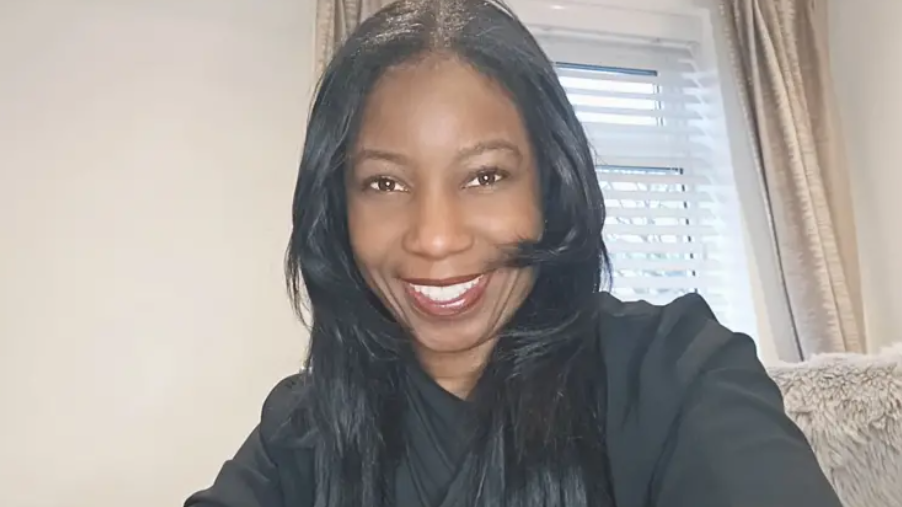 A woman smiles at the camera. She has long black hair and is wearing a black top, behind her is a cream wall and a window, with cream curtains