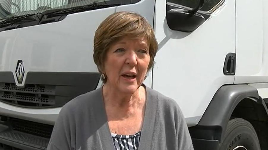 Jane Birch with short brown hair and square earrings, wearing a grey cardigan and black and white top. There is a large white lorry behind her.