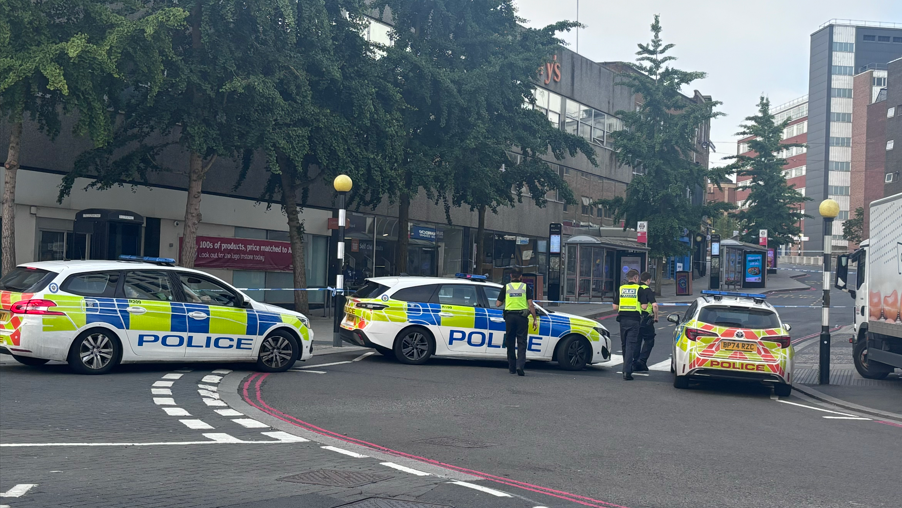 A cordoned off street with police cars and tape sectioning off a road. There are closed shops on either side of the road in the background. There are also some trees on the left side pavement in the distance.