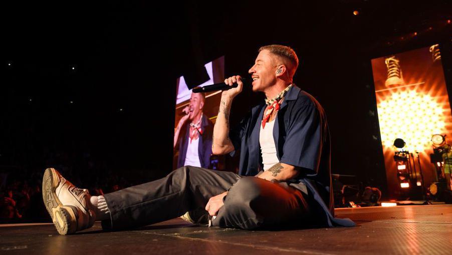 Macklemore sits down on stage, smiling, holding a microphone up to his face. There is also an image of him on a screen behind him