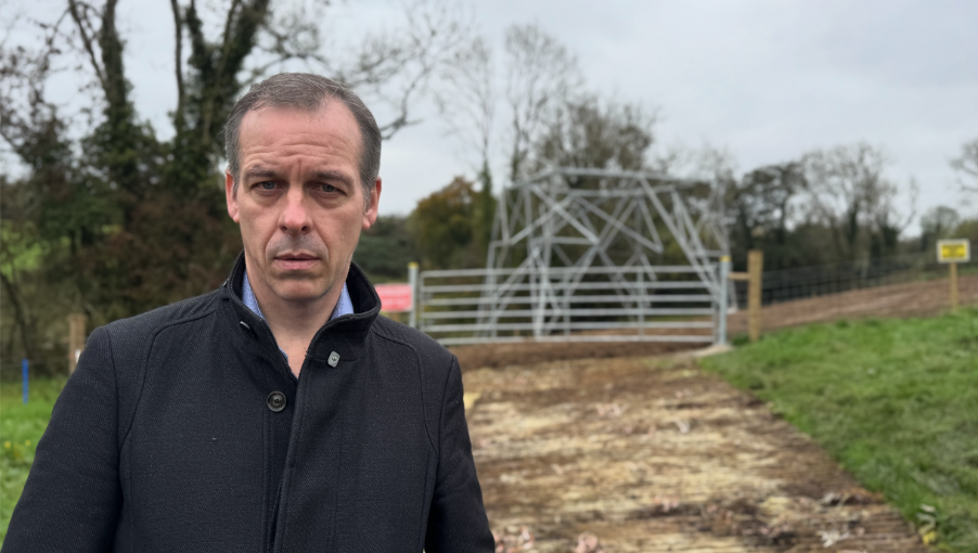 A man in a dark coat looks into the camera lens, behind him is a dirt track leading to a partially constructed electricity pylon. The track is flanked by green grass and trees, ending at an aluminium gate and wire fencing bearing warning signs. 