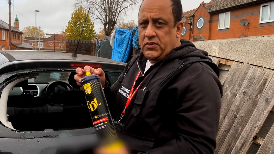 A man looking into the camera holding a nitrous oxide canister. There is a smashed car behind him. 