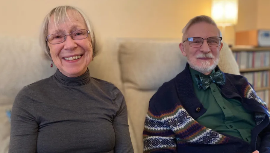 Eve and Peter Beyfus smiling while sitting on couch in home. They are both wearing glasses, Eva is wearing a grey top and Peter is wearing a bowtie with a green shirt. 