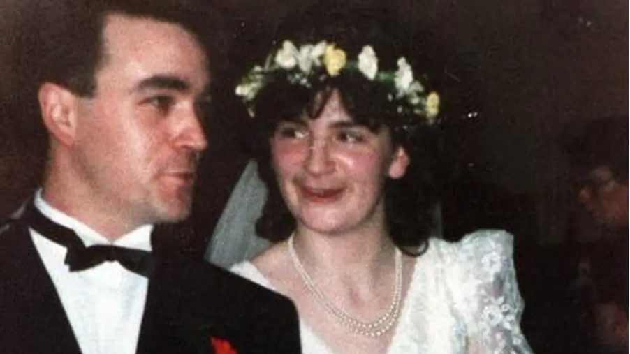 A woman in a wedding dress, peal necklace, and a flower crown. Beside her a man wearing a suit and bow tie