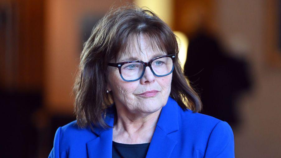 Jeane Freeman, who has brown hair and black-rimmed glasses, is wearing a blue jacket with a black top
