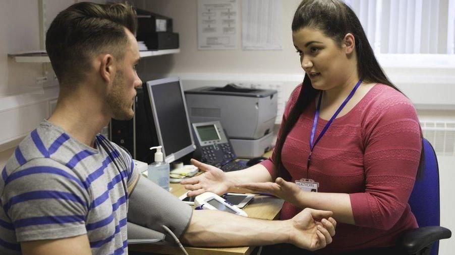 A patient, wearing a blood pressure monitor on arm, getting assessed by a medical professional