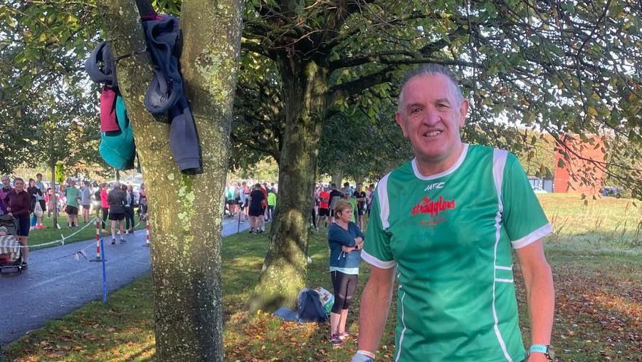 John Deery is standing in Ormeau Park in Belfast, where a Parkrun is held. He is wearing a green short-sleeved running top and is smiling at the camera. He is standing beside a tree which has some tops in between the branches. There are lots of runners off to the right hand side.