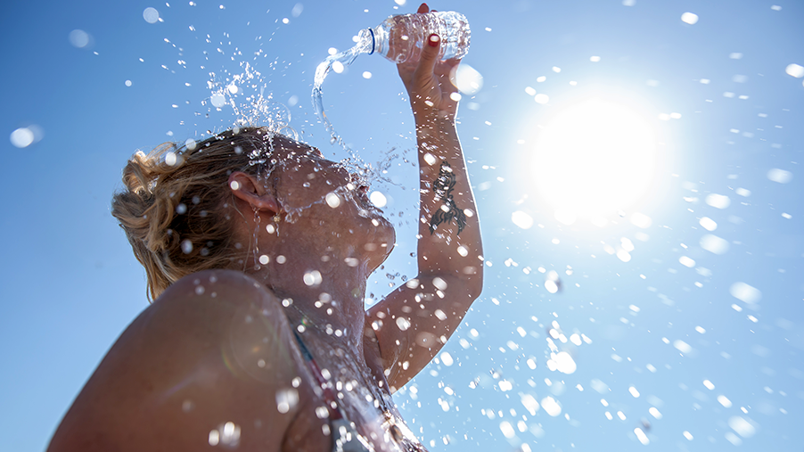 A woman pours water on herself from a plastic bottle as the sun beats down from a clear blue sky.