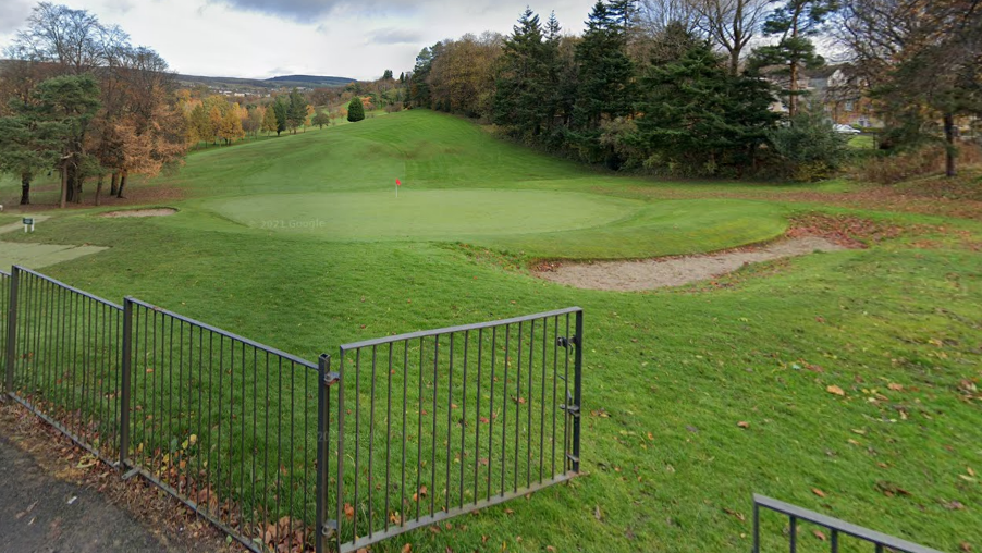 A general view of the tee at Dalmuir Golf Course. There is a grey fence in the foreground of the image. On the tee, a golfer in dark clothing is swinging a club. He is surrounded by greenery on the course.