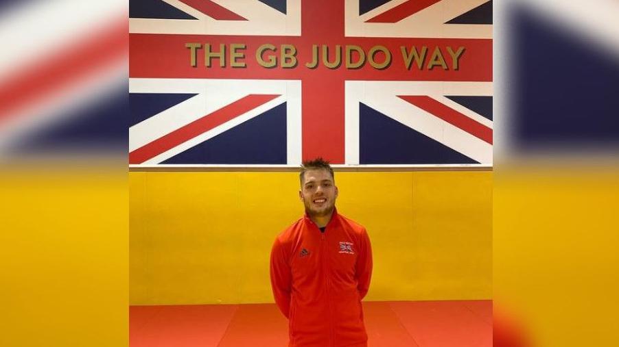 Mr Kinsella stands in front of a Union Jack which has 'THE GB JUDO WAY' written on it. He is standing with his arms behind his back and is smiling at the camera. He is wearing a red, long-sleeve jumper with the Great Britain team logo on it.