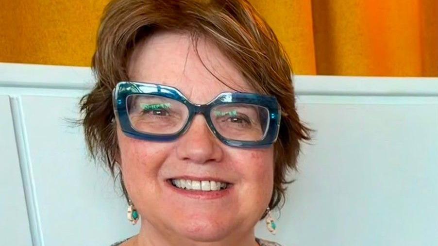 The photo shows a person with short brown hair wearing large blue-framed glasses. They are dressed in a light blue patterned blouse paired with a sheer lavender cardigan. The background features a pale-blue wall panel and a yellow curtain. The lighting is soft and natural, giving the scene a relaxed feel. 