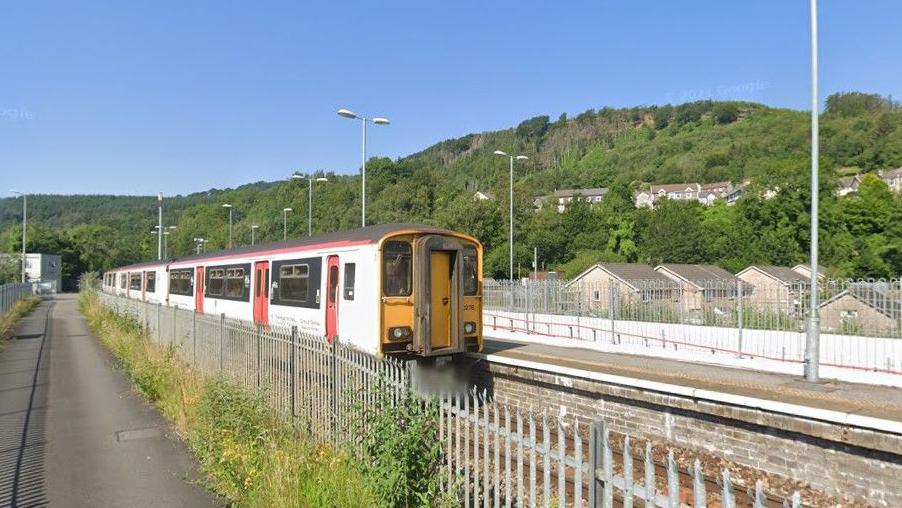 Abercynon train station. A white train with red doors is pulled up at the platform, with a metal fence on the other side of the tracks. A hillside covered in trees can be seen behind it.