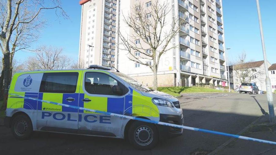 A police van sits behind blue and white police tape which is cordoning off a block of flats