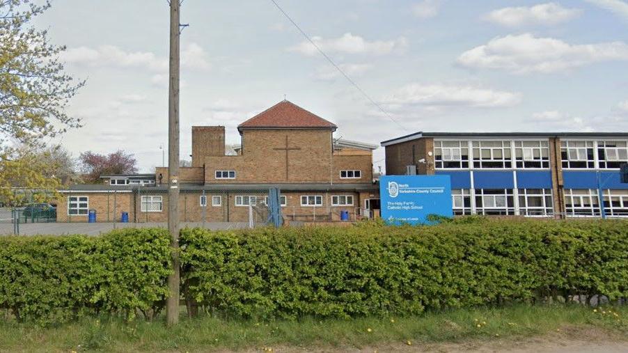 The outside of a school with brutalist architecture. It is pictured from behind a hedgerow. A cross on the outside of one of the buildings denotes the school's Catholic heritage.