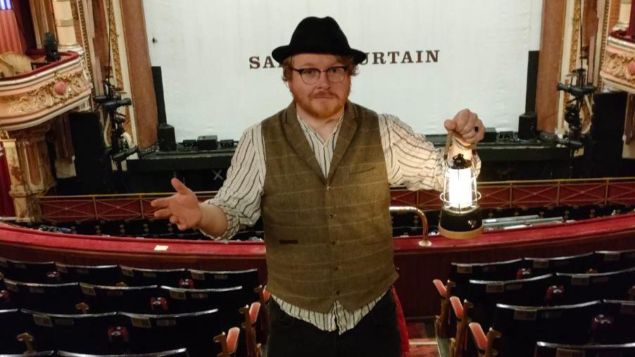 Mark has red hair, a moustache, beard and glasses. He is wearing a waistcoat, while standing on the stairs at one of the higher galleries in the King's with the stage behind him. He is holding a lamp. 