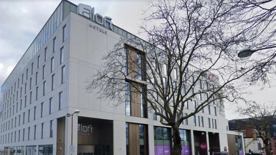 An exterior shot of the Unite hotel and conference centre in Birmingham. It is a grey building with a foyer with a sign above the door which bears the name of the Aloft hotel chain which is based there.