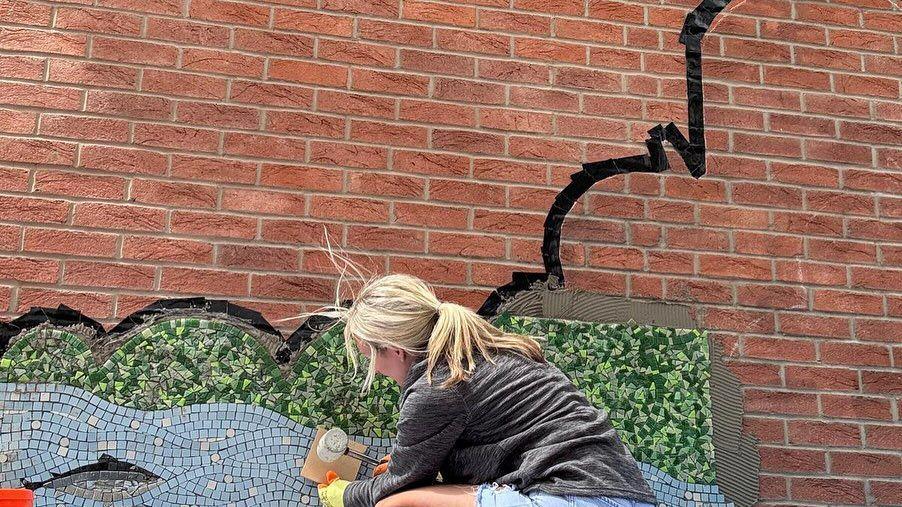 A lady with long blonde hair and a grey top has her back to the camera. She is working on the part-completed mosaic.