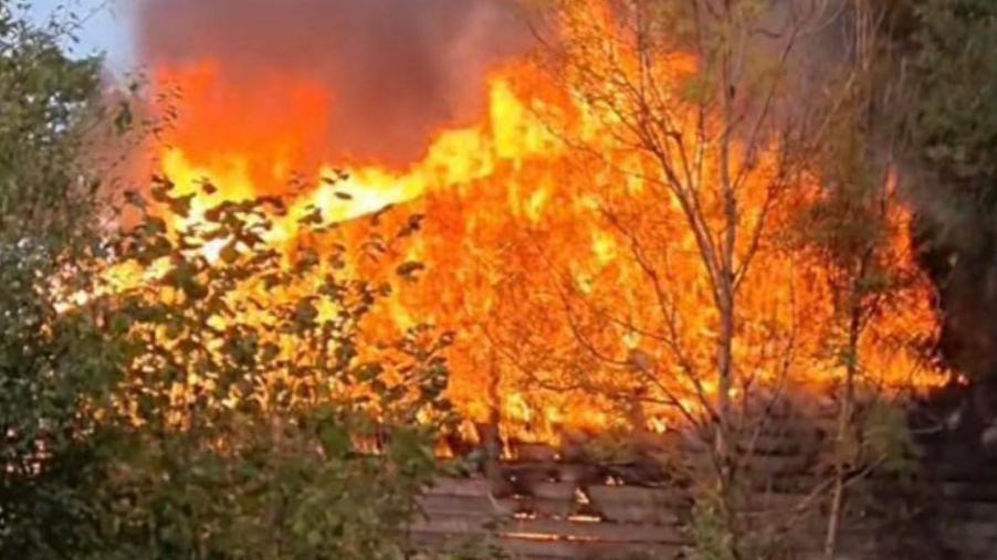 A large section of the roof on fire. The fire is orange and red and there are trees in front of it.