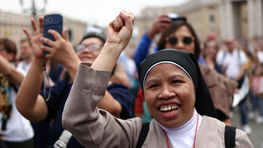 A nun in brown coat and black and white headwear with a crowd behind her