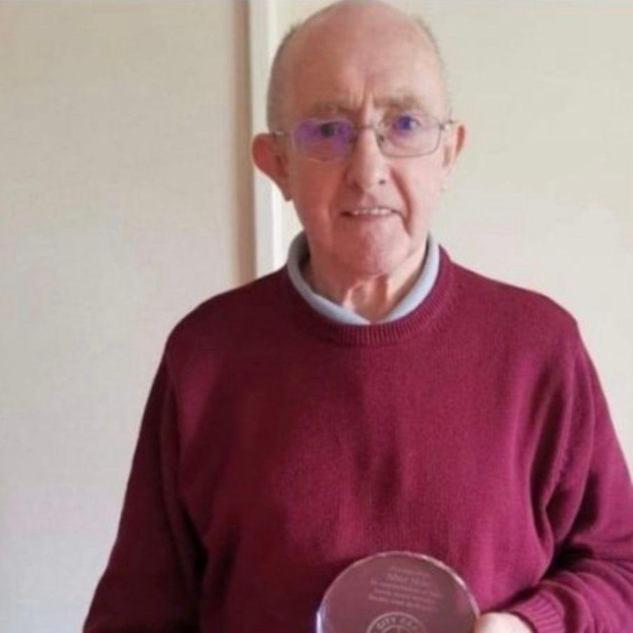 Allan Moir wearing glasses and a maroon jumper with a light-coloured top underneath it. He is holding a circular glass object.