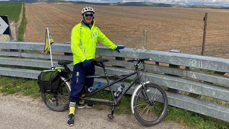 James Shone stands astride a tandem bicycle in front of a field. 