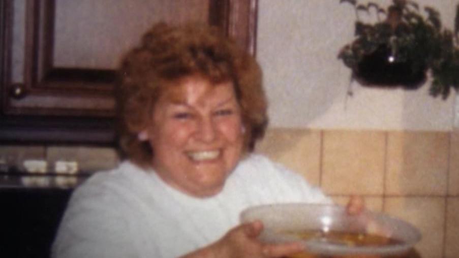 Nora Tait is smiling at the camera, she has light brown hair and a big smile on her face, She is wearing a white jumper and holding a bowl up to the camera