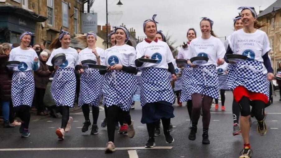 A group of eleven women can be seen running each wearing a skirt, blue and white checked apron and headscarfs. They are all carrying a frying pan. 