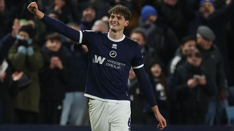 Caleb Taylor celebrates after scoring for Millwall, with his right arm raised and his hand clenched in a fist while smiling, facing away from the Millwall fans and into the pitch