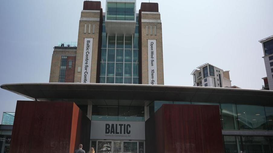 A front view of the Baltic - the glass lift is visible with the venue's opening times emblazoned on the side. Below a roof is the entrance with two red, box-shaped pillars and two people can be seen going inside.