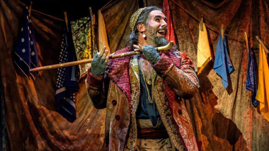 Smiling Simon Lipkin as Fagin on stage dressed in garish coloured layers and holding a cane.