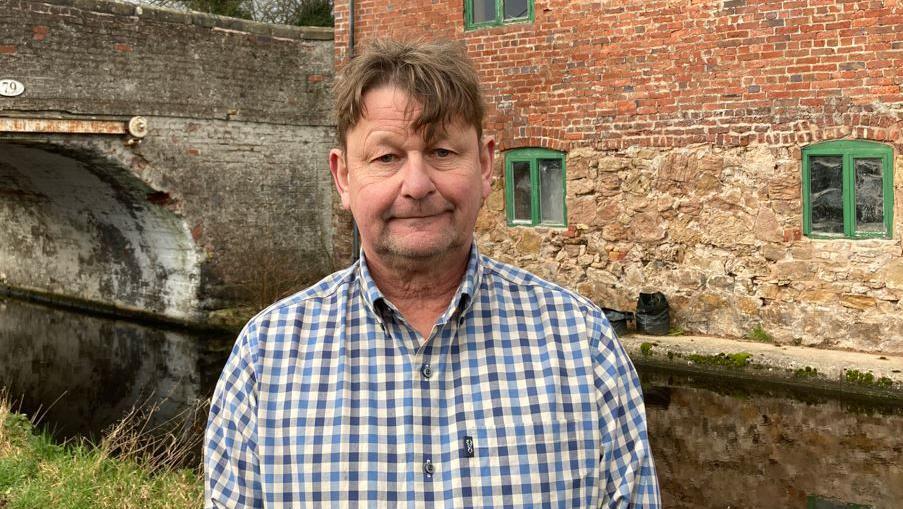 A man in a blue and white chequed shirt with light brown hair, standing in front of a canal with a stone building and a stone bridge behind him. The stone building has green-framed windows.