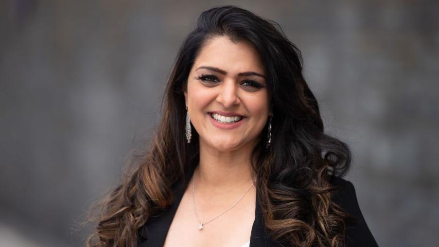 Natasha Asghar photographed, she has brown long curly hair and smiles at the