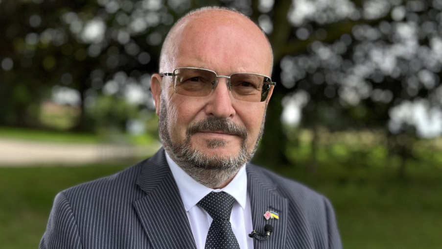 Graham Plant in tinted-lens spectacles, a pin-striped dark jacket, white shirt and black tie with tiny white spots 